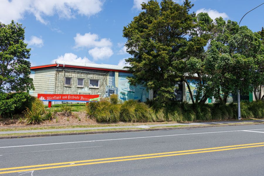 A low building behind trees and flax, seen across Port Albert Road in Wellsford.