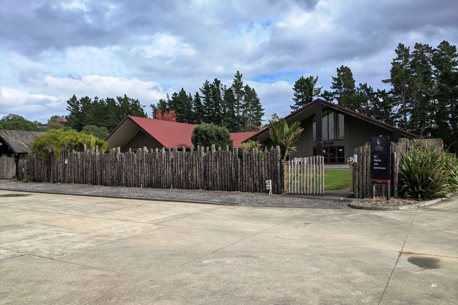 A palisade fence and buildings with red roofs at Te Hana Te Ao Marama, under a cloudy sky.